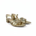 A pair of metallic gold strappy sandals with low block heels and pointed toes, shown on a light gray background.