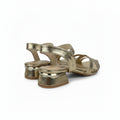 A pair of metallic gold strappy slingback sandals with low block heels, shown on a light gray fabric studio background.