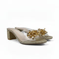 A pair of women's heeled mule sandals with clear transparent straps, a chunky glittery gold block heel, and a decorative gold beaded floral embellishment across the toe area, shown on a plain white background.