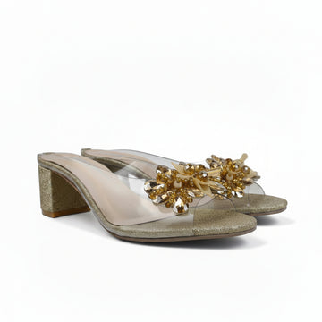 A pair of women's heeled mule sandals with clear transparent straps, a chunky glittery gold block heel, and a decorative gold beaded floral embellishment across the toe area, shown on a plain white background.