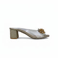 A single women's heeled mule sandal shown in profile against a solid white background. The shoe features a glittery gold block heel, a clear transparent upper, and a decorative gold floral embellishment on the toe strap.