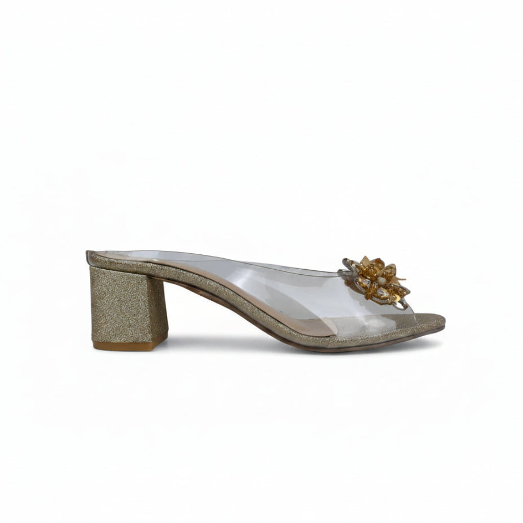 A single women's heeled mule sandal shown in profile against a solid white background. The shoe features a glittery gold block heel, a clear transparent upper, and a decorative gold floral embellishment on the toe strap.