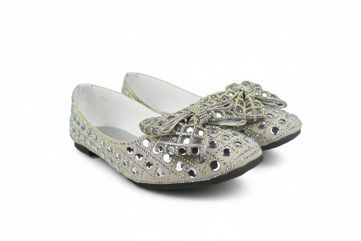 A pair of silver glitter flat shoes embellished with rhinestones and a large decorative bow on the toe, shown against a plain white background.