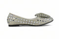 A single women's flat shoe in a light champagne or silver-gray suede, heavily embellished with rhinestones and mirrored studs, featuring a matching embellished fabric bow on the toe, set against a plain white background.