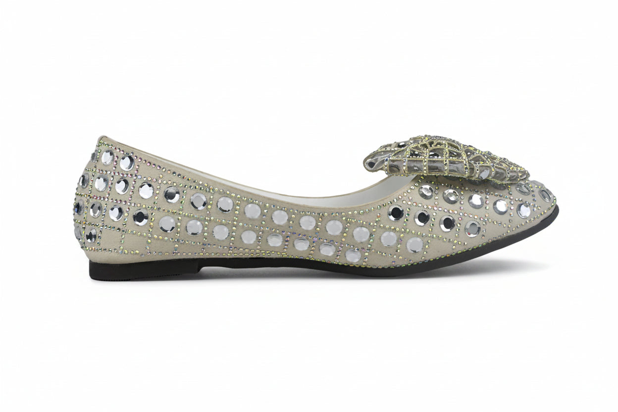 A single women's flat shoe in a light champagne or silver-gray suede, heavily embellished with rhinestones and mirrored studs, featuring a matching embellished fabric bow on the toe, set against a plain white background.