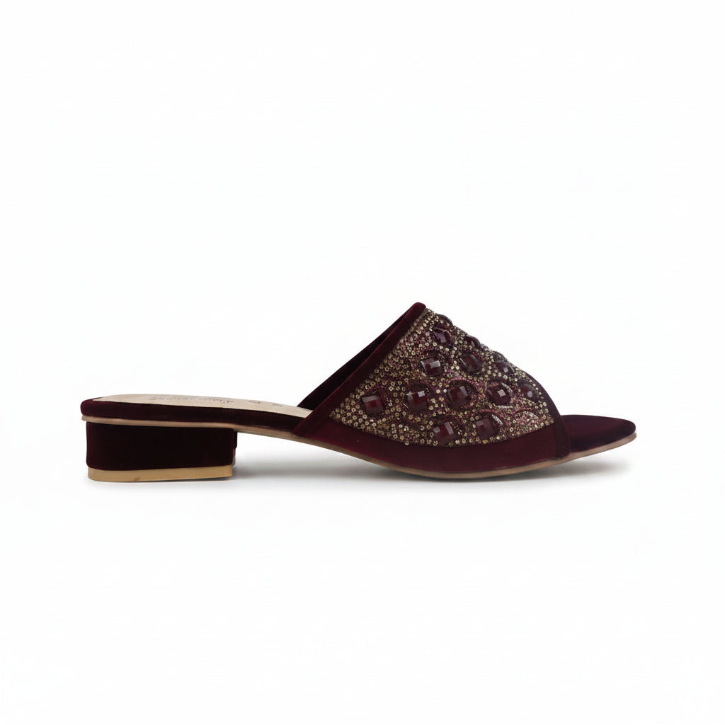 A single burgundy velvet mule sandal with an embellished strap featuring dark red and gold rhinestone-like details, shown from a side profile against a light gray background.