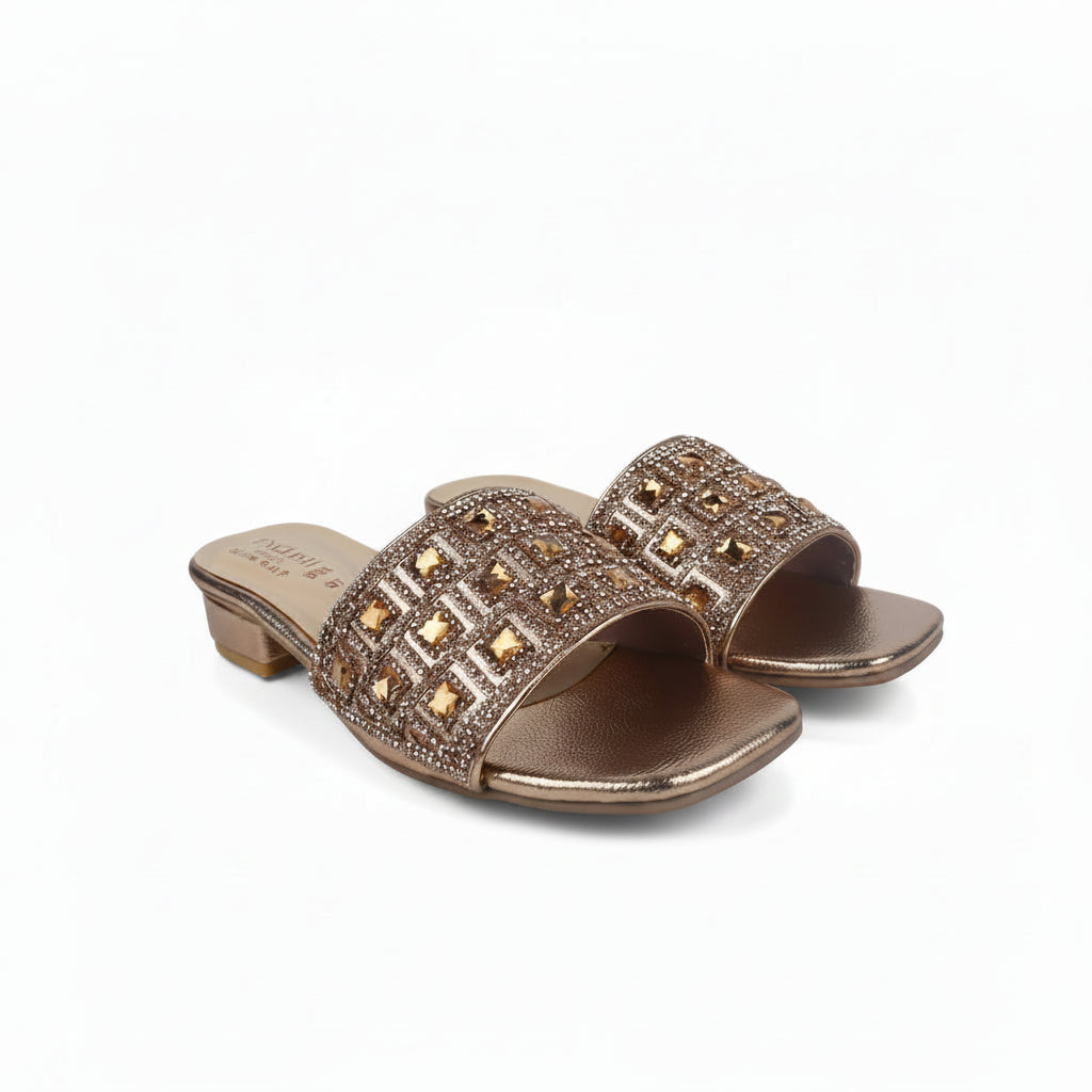 A pair of metallic rose gold slide sandals with geometric rhinestone embellishments on the straps and a low block heel, shown on a light gray background.