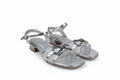 A pair of metallic silver strappy sandals with block heels and decorative metal hardware on the toes, shown against a light gray fabric backdrop.