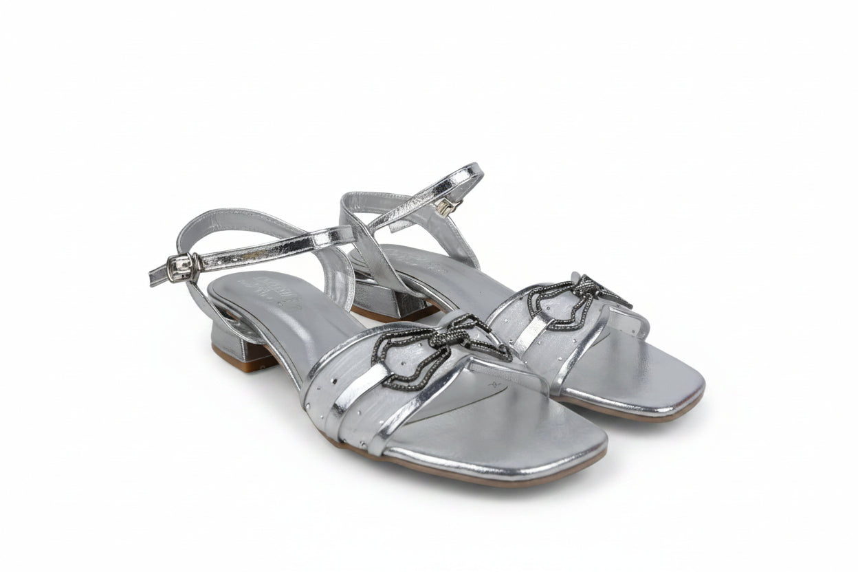 A pair of metallic silver strappy sandals with block heels and decorative metal hardware on the toes, shown against a light gray fabric backdrop.