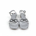 A pair of metallic silver square-toe heeled sandals with thin straps, bow-shaped embellishments, and ankle straps, photographed on a light gray surface.