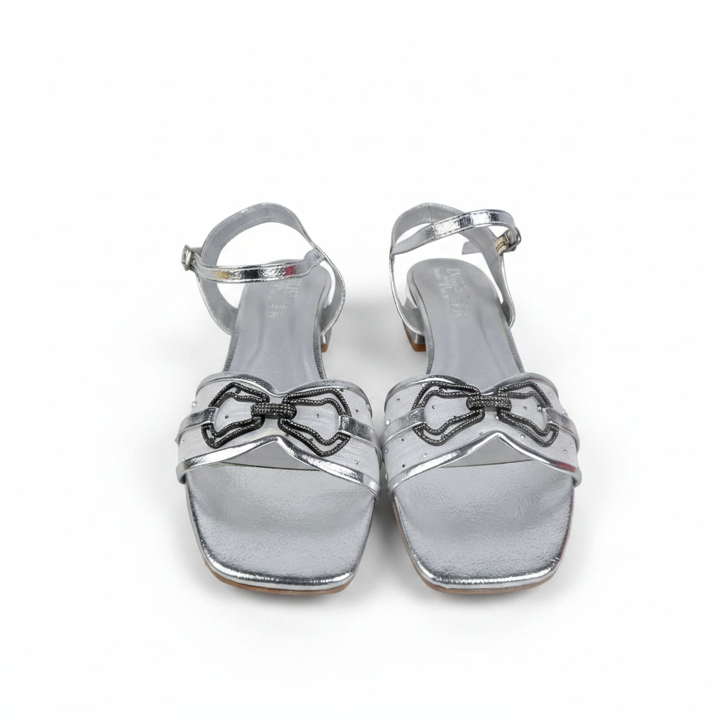 A pair of metallic silver square-toe heeled sandals with thin straps, bow-shaped embellishments, and ankle straps, photographed on a light gray surface.