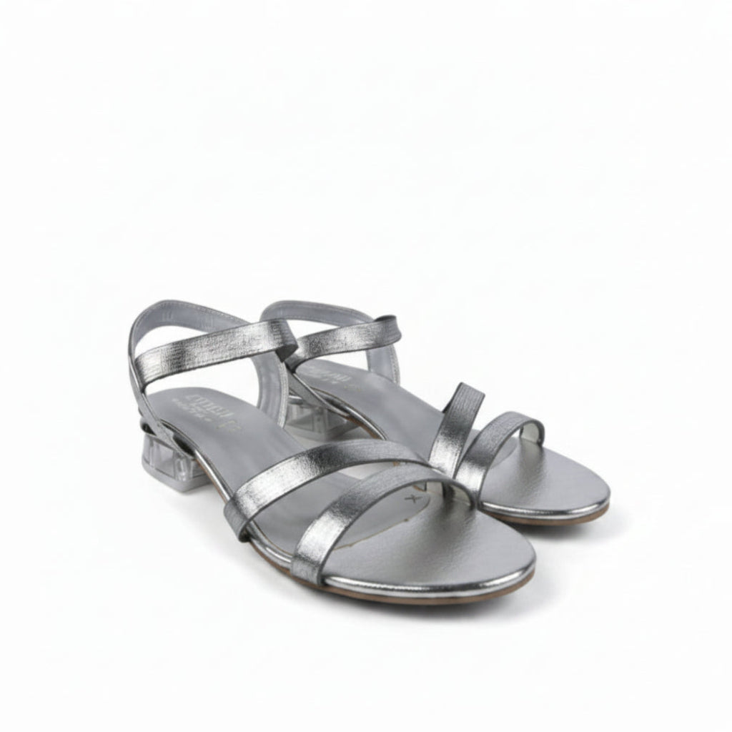 A pair of metallic silver strappy sandals with low block heels, shown on a light gray fabric background.