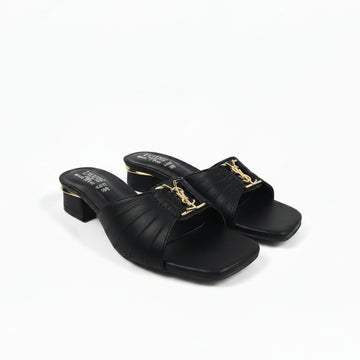 A pair of black leather slide sandals with a low block heel and a gold-tone metal YSL logo buckle, shown on a light gray background.