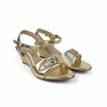 A pair of metallic gold heeled sandals with ankle straps and decorative gold hardware on the toes, photographed on a light gray fabric background.