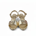A pair of gold metallic open-toe heeled sandals with ankle straps and decorative buckle details, shown on a light gray background.
