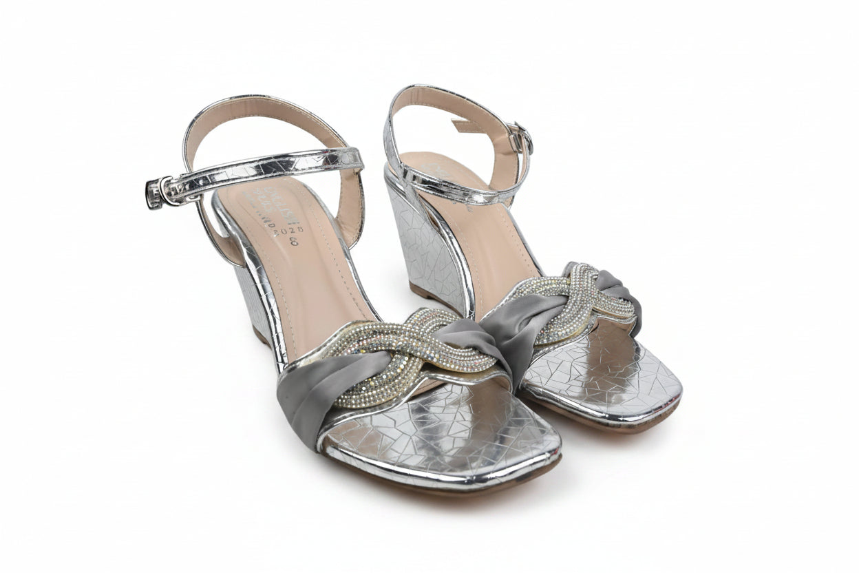 A pair of women's metallic silver wedge sandals with ankle straps, rhinestone-embellished toe straps, and a knotted bow detail on the front, shown on a white studio background.