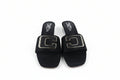 A pair of black slide sandals with large square gold-tone metal C-shaped hardware on the straps, photographed on a light gray surface.