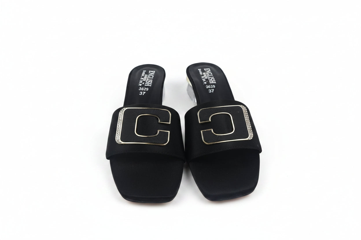 A pair of black slide sandals with large square gold-tone metal C-shaped hardware on the straps, photographed on a light gray surface.