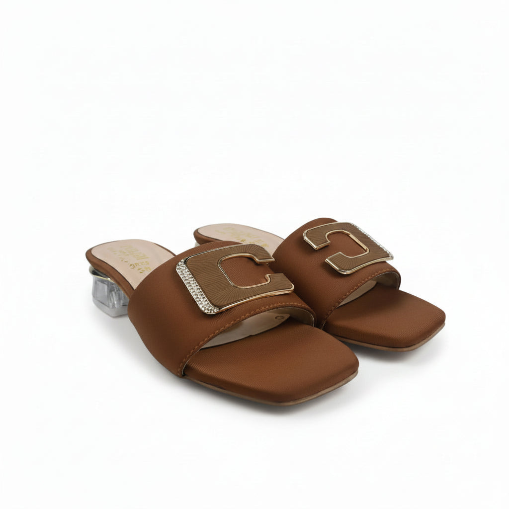 A pair of women's brown leather slide sandals with square toes and large decorative gold-tone metal buckles, displayed on a light gray fabric background.