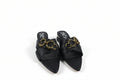 A pair of black pointed-toe mule sandals with wide crisscross straps and a prominent gold-tone chain detail across the top, shown on a light gray surface.