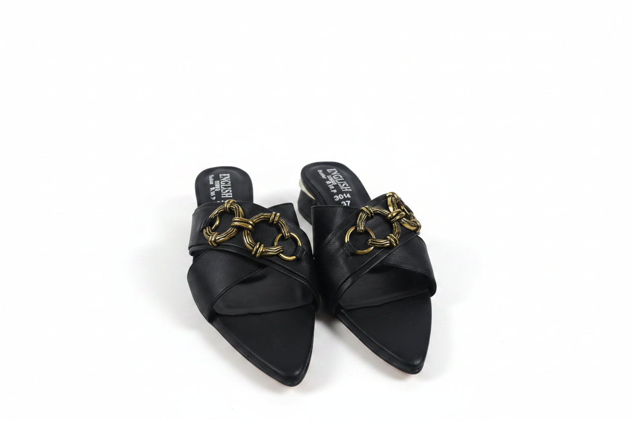 A pair of black pointed-toe mule sandals with wide crisscross straps and a prominent gold-tone chain detail across the top, shown on a light gray surface.
