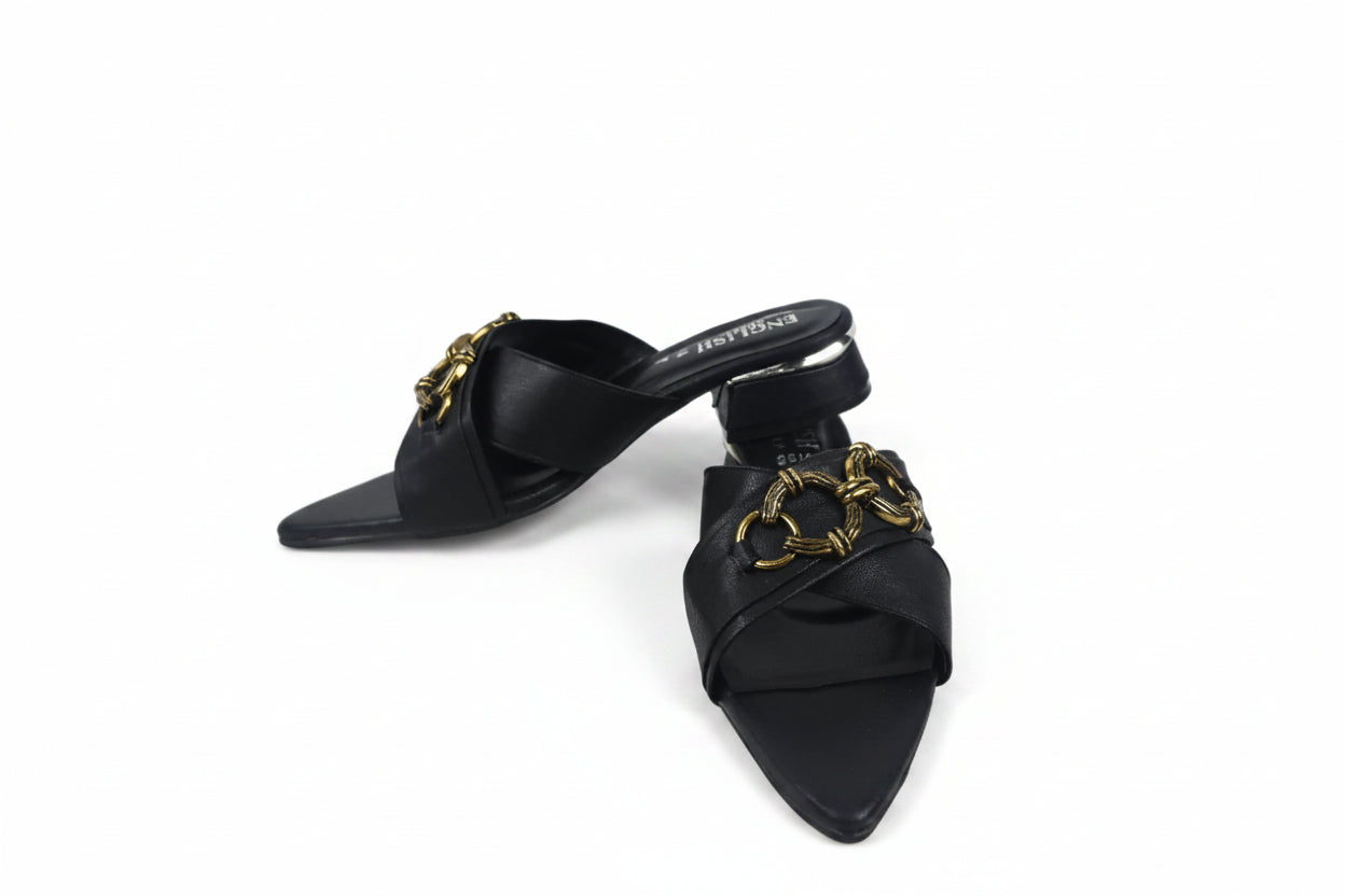 A pair of black leather pointed-toe mule sandals with chunky gold-tone chain embellishments across the straps, displayed on a light gray floor.