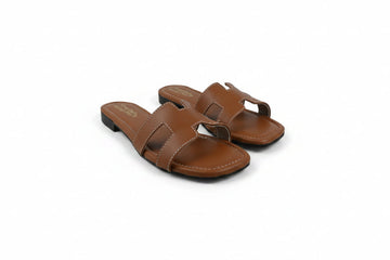A pair of women's flat slide sandals in a warm brown leather, featuring a double strap design with a small central cutout, photographed on a white studio background.