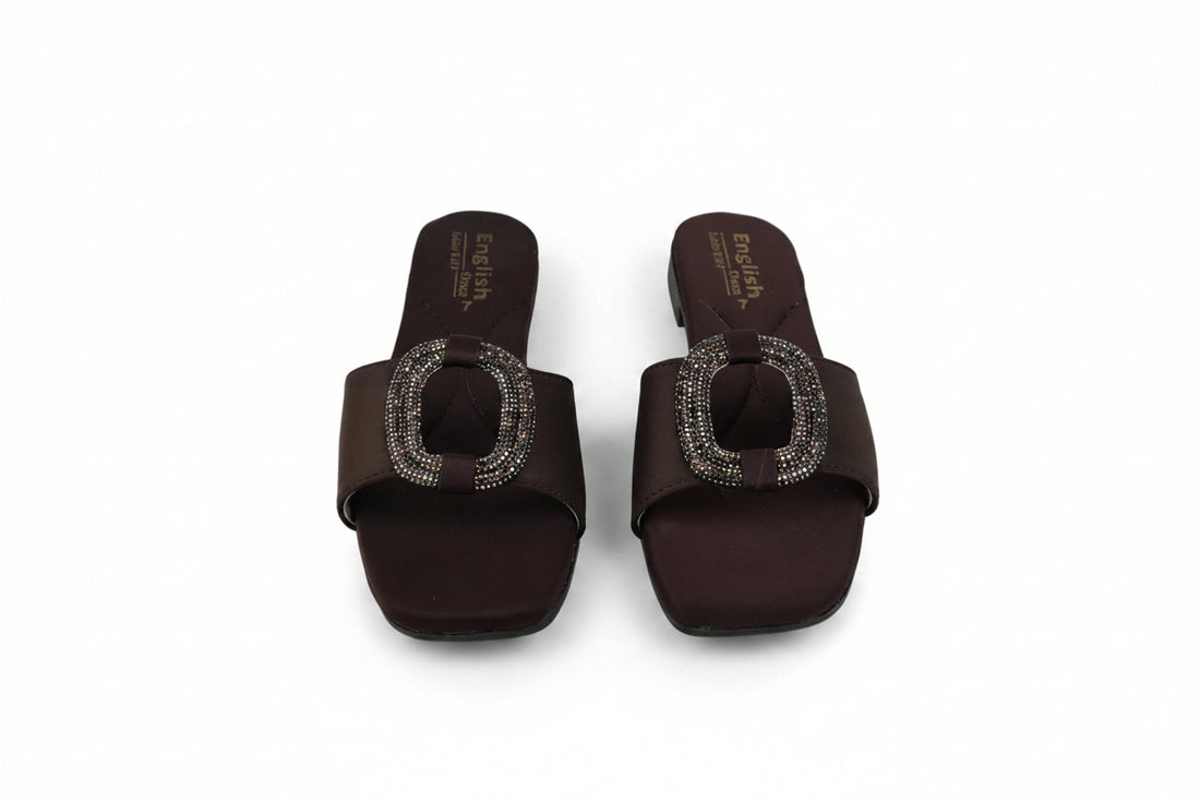 A pair of dark brown slide sandals with square toes and a prominent oval buckle embellished with sparkling rhinestones, shown on a light gray background.