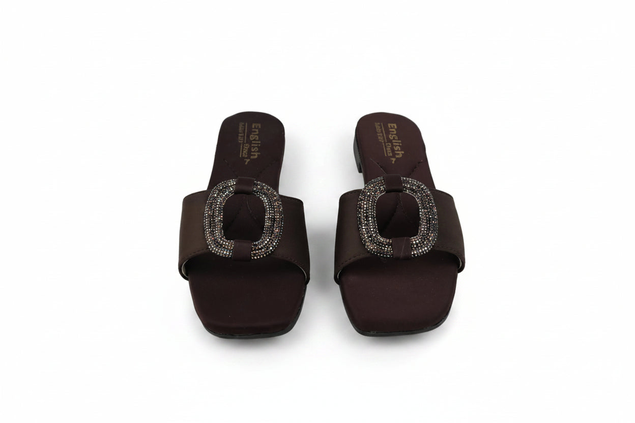 A pair of dark brown slide sandals with square toes and a prominent oval buckle embellished with sparkling rhinestones, shown on a light gray background.