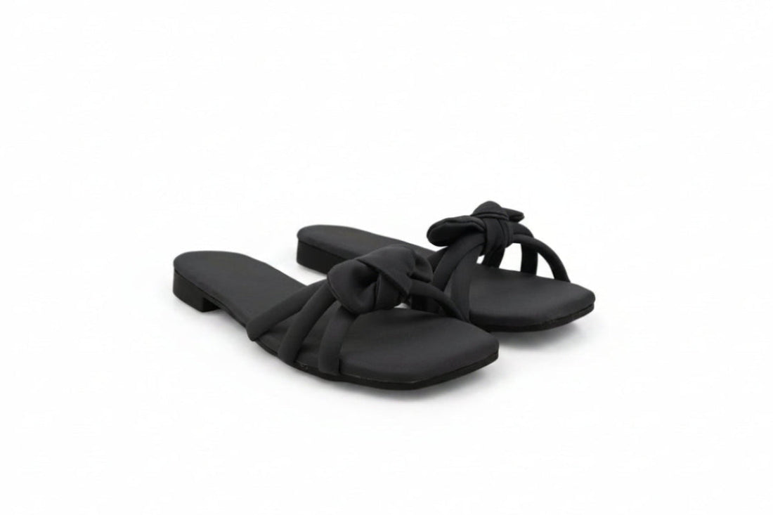 A pair of black slide sandals with knotted strap details, photographed on a light gray fabric backdrop.