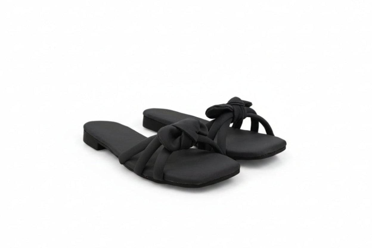 A pair of black slide sandals with knotted strap details, photographed on a light gray fabric backdrop.
