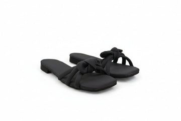 A pair of black slide sandals with knotted strap details, photographed on a light gray fabric backdrop.