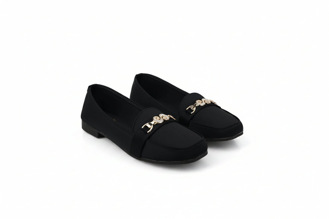 A pair of black slip-on flats with a gold-tone decorative chain link detail across the vamp, displayed on a light gray studio background.