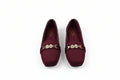 A pair of burgundy flat loafers with a gold-tone chain detail across the vamp, displayed on a light gray surface.