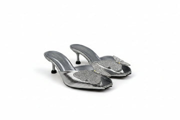 A pair of metallic silver mule heels with rhinestone-embellished toe straps, photographed on a light gray fabric backdrop.