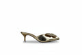 A single metallic gold mule sandal with a pointed toe, a sculptural floral embellishment on the vamp, and a slim kitten heel, shown against a neutral light gray backdrop.