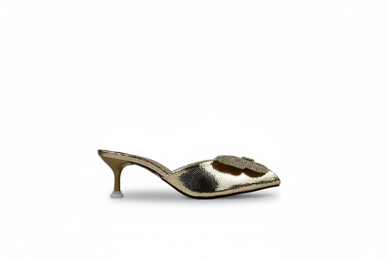 A single metallic gold mule sandal with a pointed toe, a sculptural floral embellishment on the vamp, and a slim kitten heel, shown against a neutral light gray backdrop.