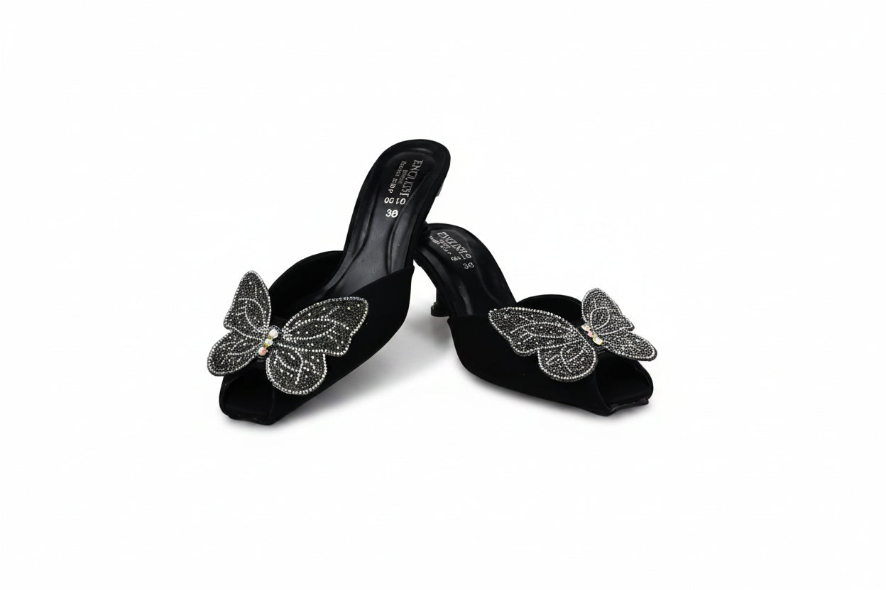 A pair of black velvet mule sandals with high heels, decorated with large silver butterfly embellishments on the front, set against a plain white backdrop.
