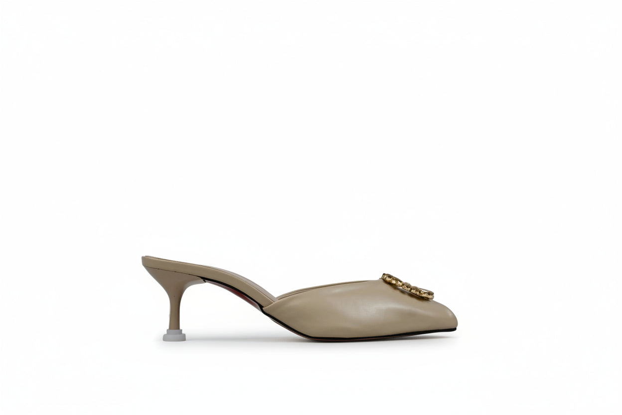 A single beige leather mule high-heel shoe with a pointed toe and a gold-tone metal logo clasp, shown in profile against a textured light gray background.