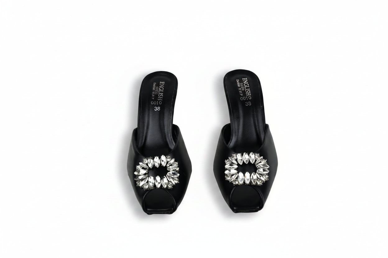 A pair of black mule sandals with clear rhinestone-embellished square toe caps, shown on a light gray surface.