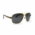 A pair of aviator-style sunglasses with a gold metal frame and dark gray lenses, displayed on a white surface with two black textured studio props in the background.