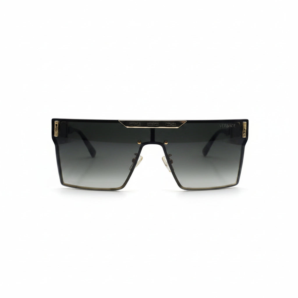 A pair of square shield sunglasses with a dark gray gradient lens and gold metal trim, displayed on a white surface with a reflective silver backdrop.