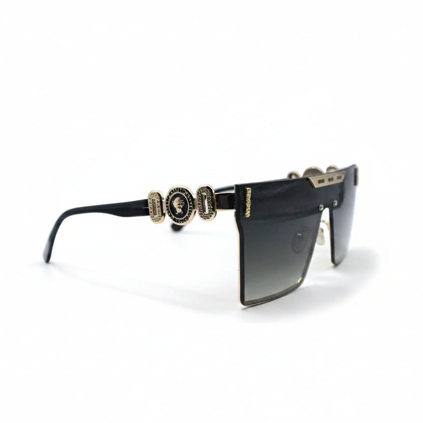 A pair of designer sunglasses with a black acetate frame, gold-tone metal accents, and gradient lenses, displayed on a white surface between two silver studio reflectors.