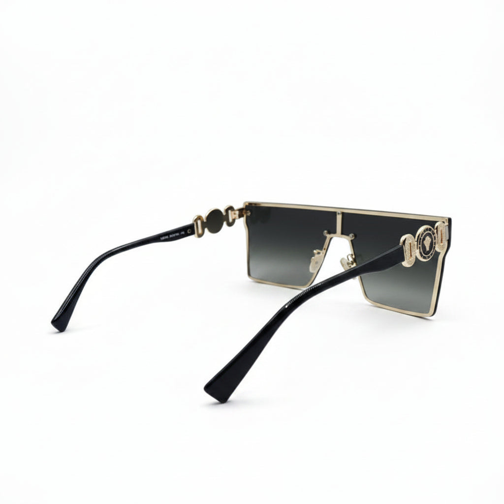 A pair of square-frame sunglasses with black temples, gold metal rims, and gradient dark lenses, displayed on a white surface with a reflective silver backdrop.