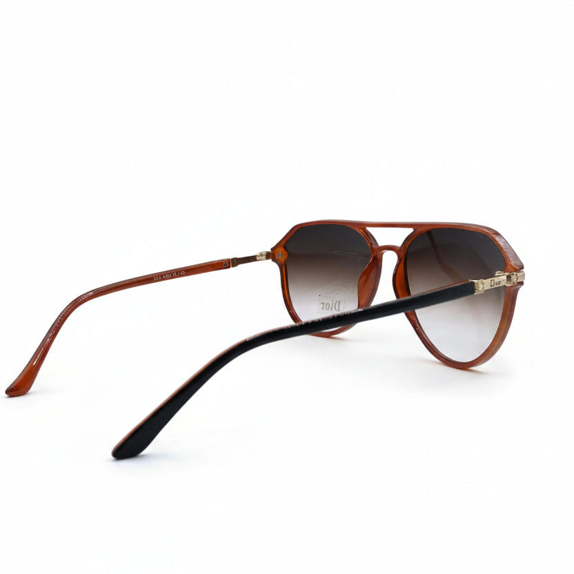 A pair of aviator-style sunglasses with a translucent brown frame, gradient brown lenses, and black temple arms, displayed on a white surface between two textured silver fabric panels.