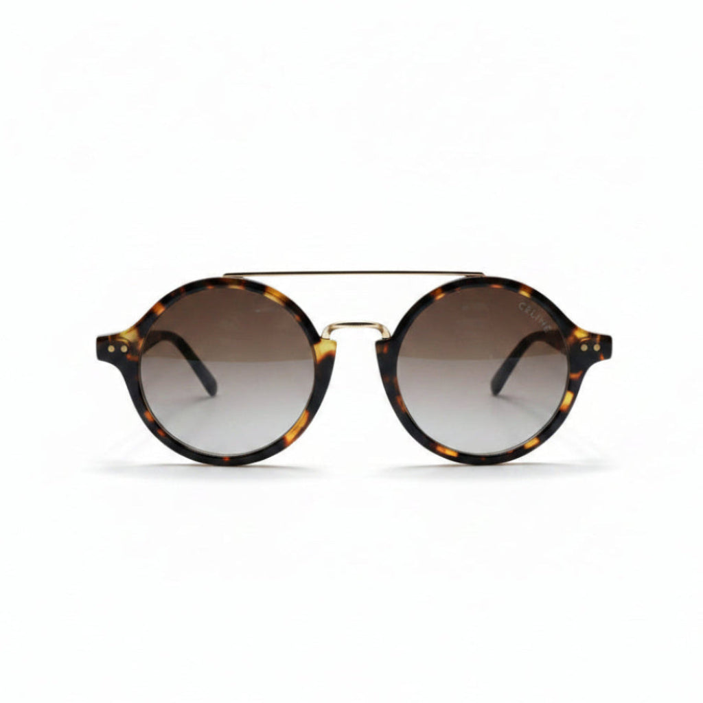 Round sunglasses with a tortoiseshell pattern, a thin gold metal bridge, and gradient brown lenses, displayed on a white surface between two silver studio reflectors.