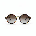 Round sunglasses with a tortoiseshell pattern, a thin gold metal bridge, and gradient brown lenses, displayed on a white surface between two silver studio reflectors.
