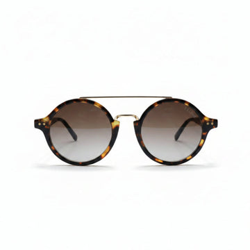Round sunglasses with a tortoiseshell pattern, a thin gold metal bridge, and gradient brown lenses, displayed on a white surface between two silver studio reflectors.