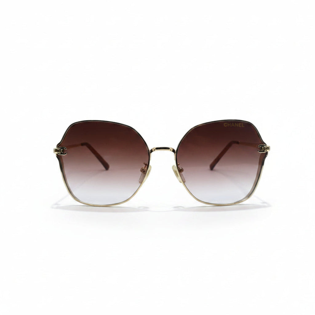 Oversized square sunglasses with a gold metal frame and brown gradient lenses, displayed on a white surface with a reflective silver backdrop.