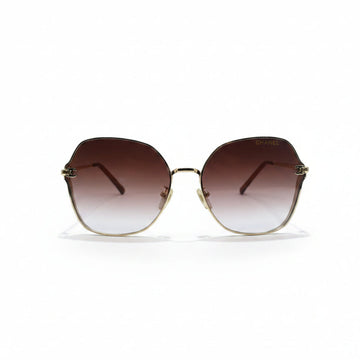 Oversized square sunglasses with a gold metal frame and brown gradient lenses, displayed on a white surface with a reflective silver backdrop.